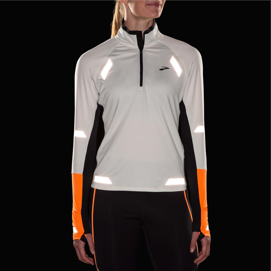 Front view of a model wearing the Brooks Women's Run Visible 1/2 Zip 2.0 in the Ecru/Fluoro Flash/Black colourway. Reflective strips are reflecting artificial light. Model is also wearing Brooks tights. (8499447333026)