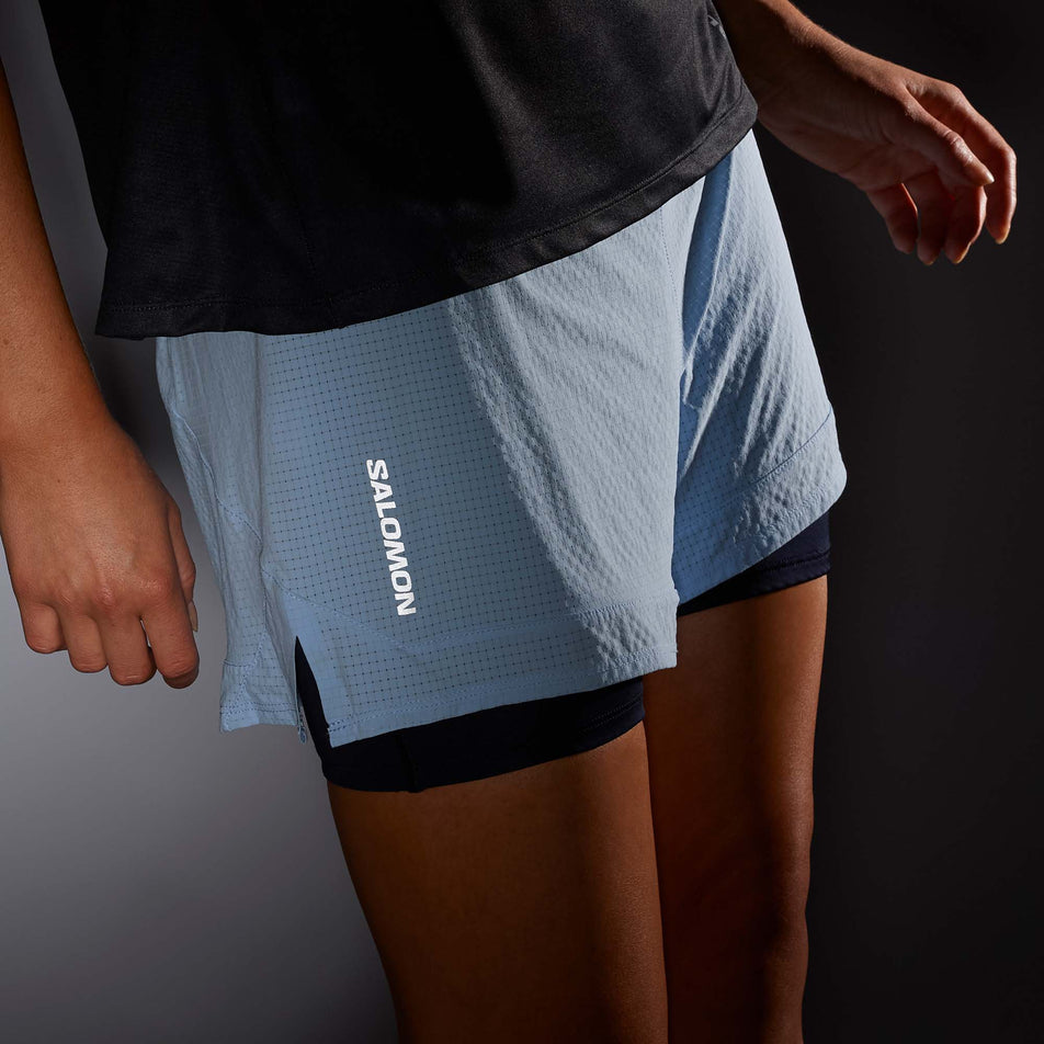 Close-up front view of a model wearing a pair of Salomon Women's Sense Aero 2in1 Shorts in low light conditions - to show the reflective Salomon logo. Chambray Blue/Deep Black colourway (7892306952354)