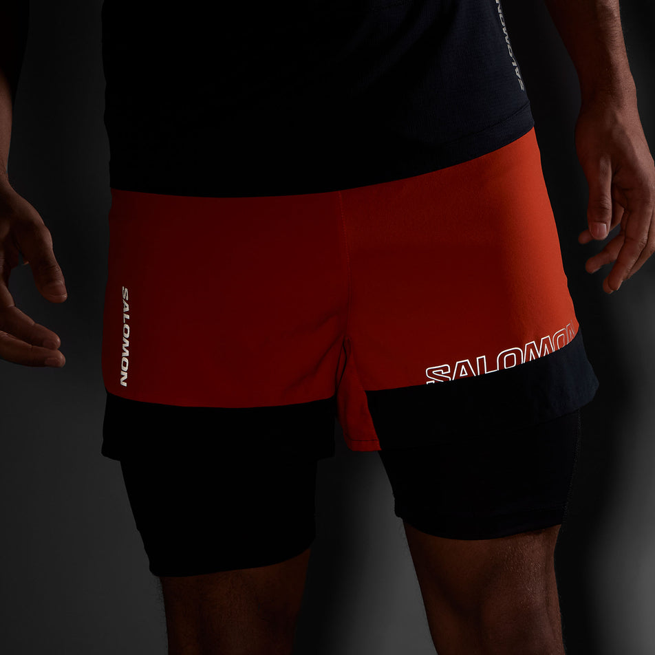 The reflective Salomon logos on the front of a pair of Salomon Men's Cross 2in1 Shorts. Shorts are being worn by a model.  (8467295436962)
