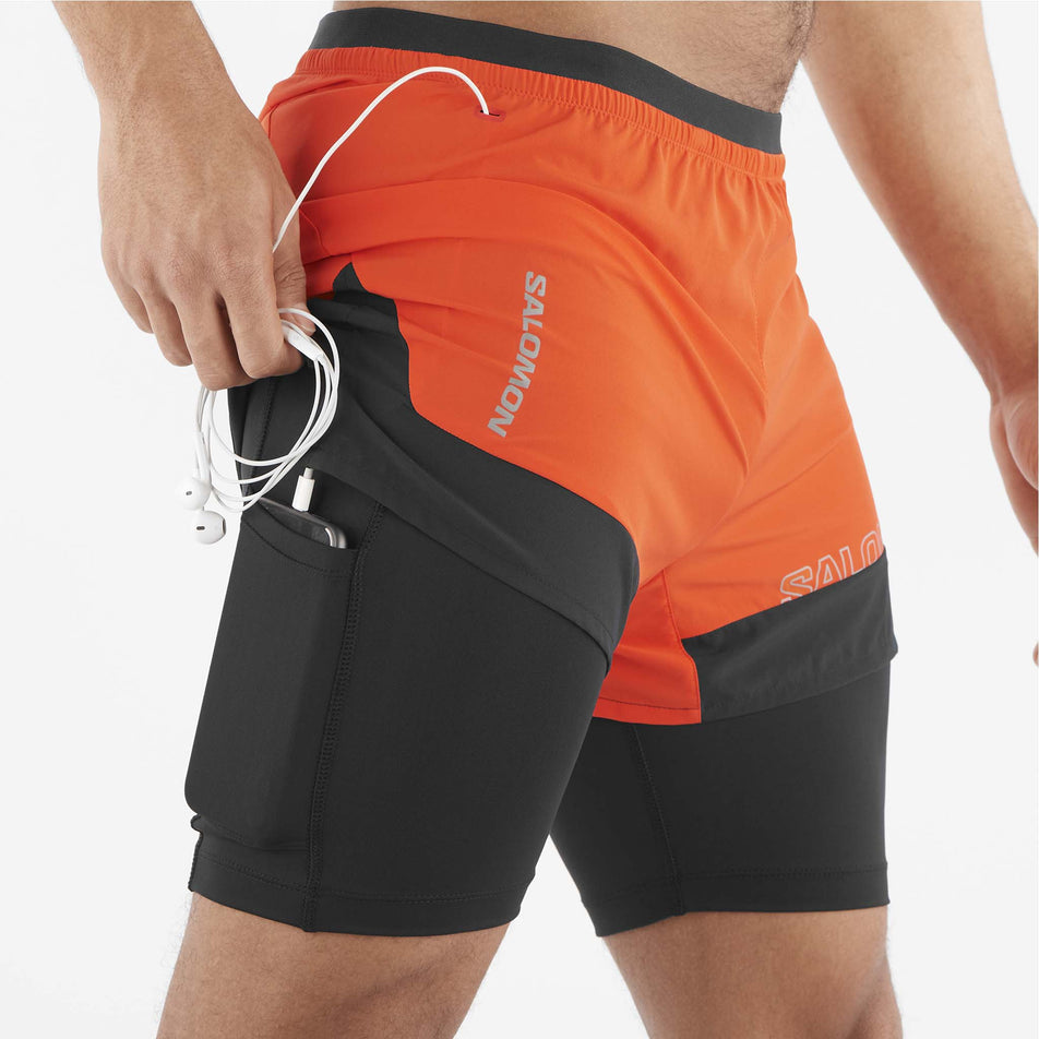 A model demonstrating that a phone can be stored in the stretch pocket located on the outside of the left-inner liner of a pair of Salomon Men's Cross 2in1 Shorts. (8467295436962)