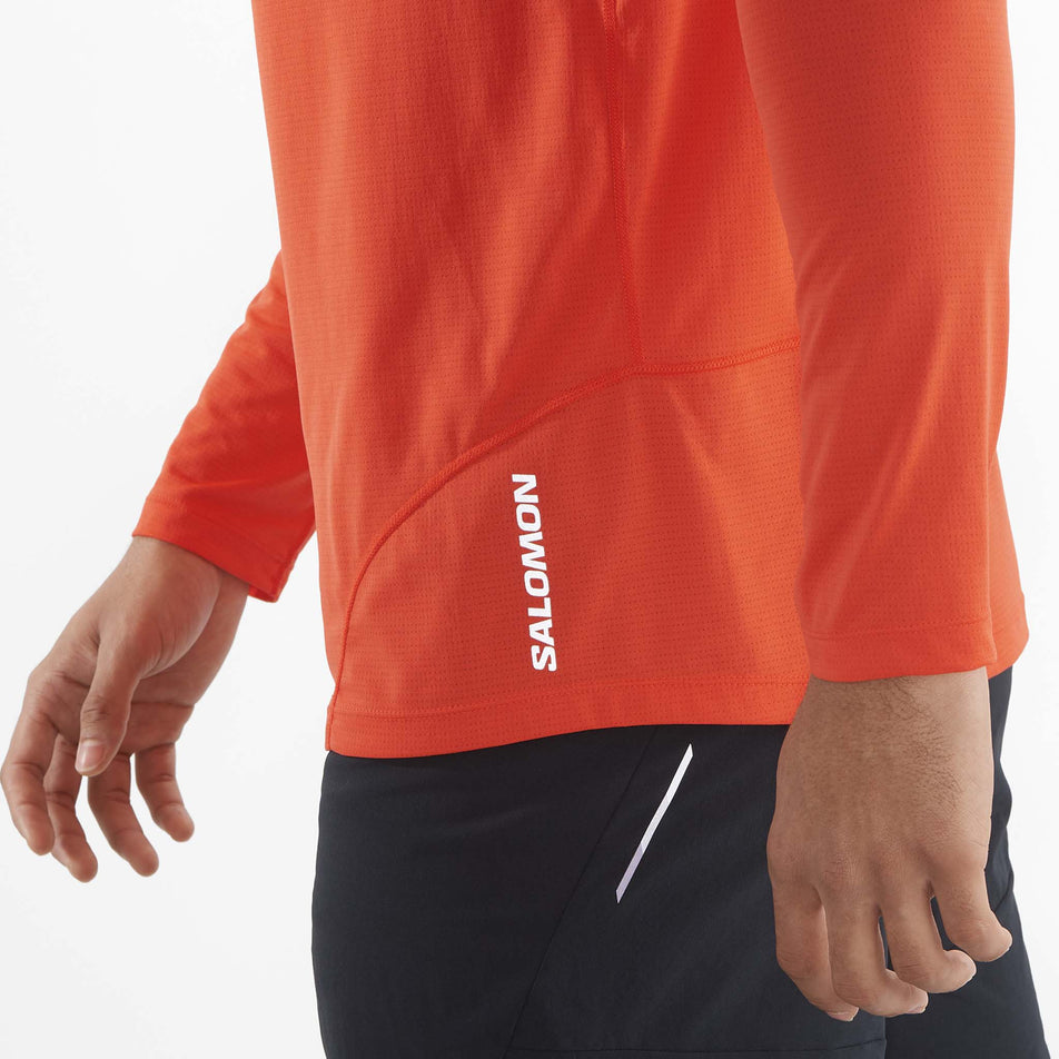 Close-up view of the reflective Salomon logo which is located on the lower front left side of a Salomon Men's Cross Run Long Sleeve T-Shirt. Top is being worn by a model. (8467287179426)