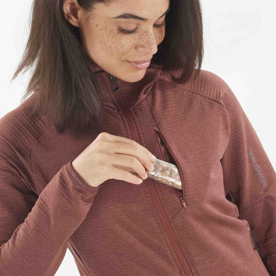 A model demonstrating that nutrition can be stored in the chest pocket of the Salomon Women's Essential Lightwarm Midlayer Jacket Hoodie. Top is being worn by a model. (8509527097506)