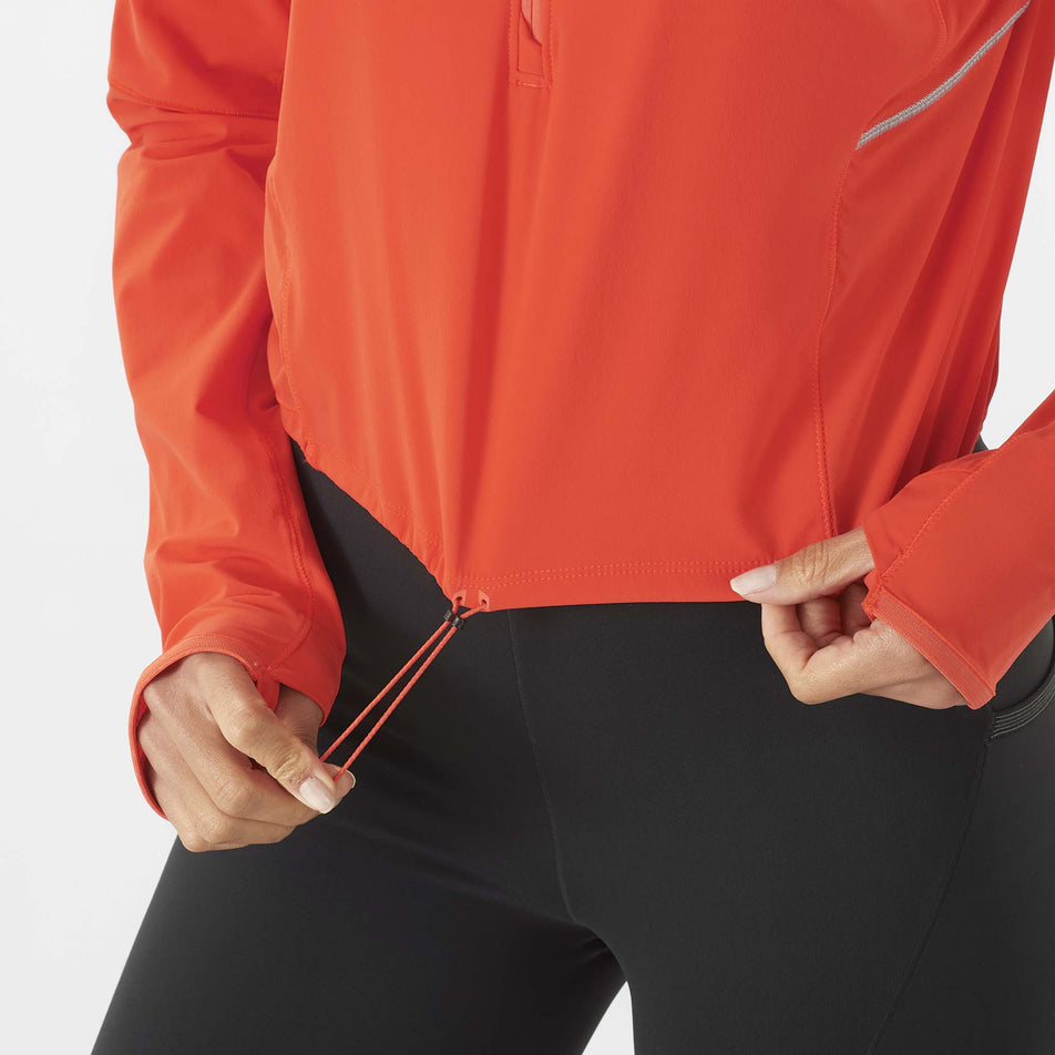 A model highlighting the drawcord on the lower hem of a Salomon Women's Sense Aero Hybrid Cropped Half Zip Midlayer in the Cherry Tomato colourway. Top is being worn by a model who is also wearing leggings. (8462927102114)